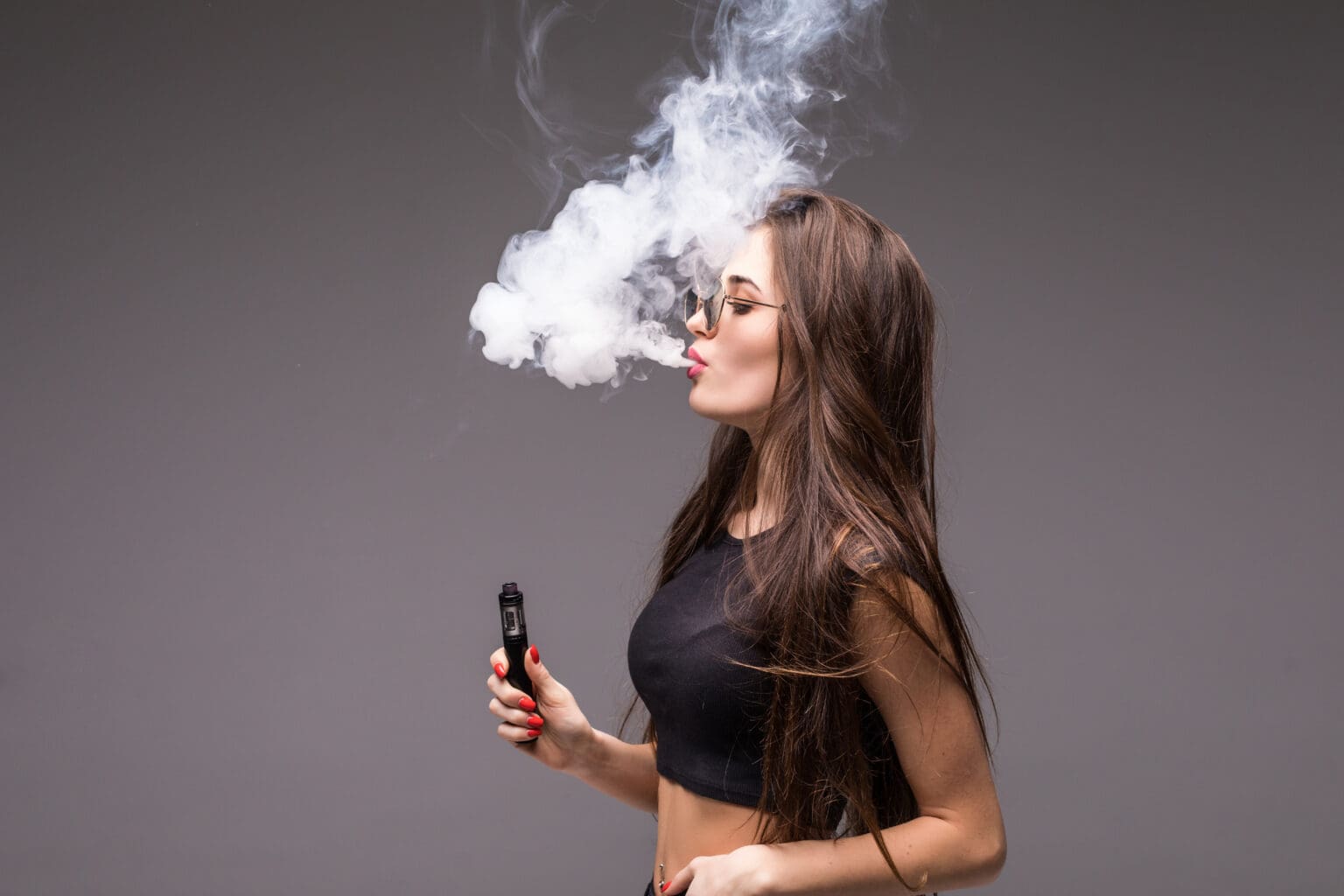 young woman sunglasses black clothes vaping blowing smoke electronic cigarette gray wall